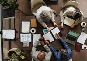 Group of middle aged multiethnic business professionals collaborating around table, reviewing documents and using laptop, top view showing teamwork and corporate meeting environment